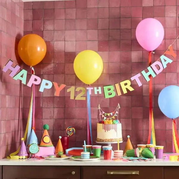 Bright birthday party setup with colourful balloons, a Happy 12th Birthday banner, striped cake, party hats, and vibrant tableware on a pink tiled background